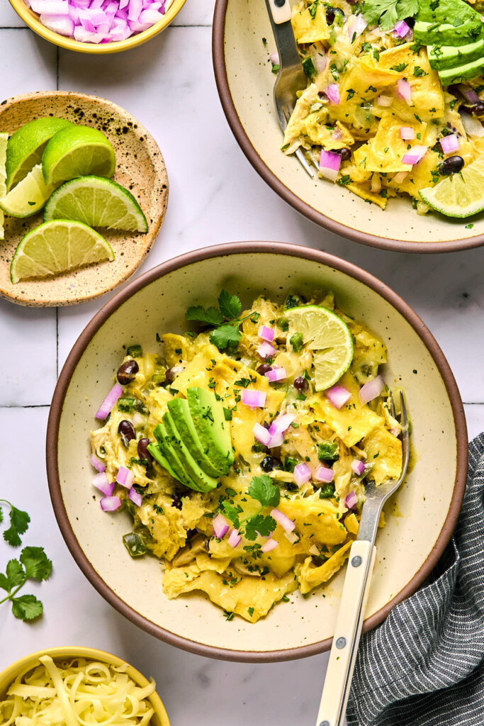 Two bowls with mixture of shredded chicken, black beans, and veggies topped with melted cheese. They are garnished with sliced avocado, diced red onions, cilantro, and a lime wedge and each bowl has a fork in it. Next to the pan is a plate of lime wedges, a bowl of diced red onion, shredded cheese, and a blue towel.
