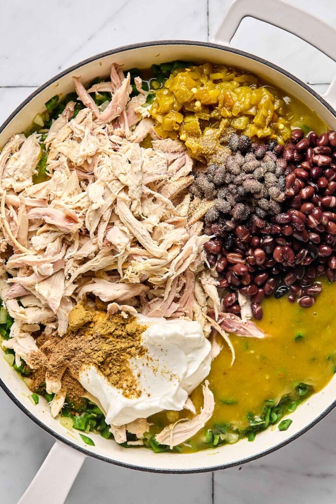 Large white pan with shredded chicken, green chiles, black beans, green enchilada sauce, greek yogurt, and spices in separate piles overtop sautéed peppers and onions.