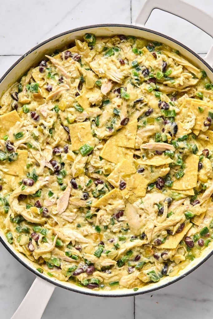 Large while skillet filled with a mixture of shredded chicken, veggies, black beans, and pieces of corn tortillas in a creamy green sauce.