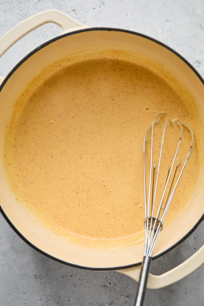 Healthy mac and cheese sauce in a large white dutch oven. There is a metal whisk in the pot as well.