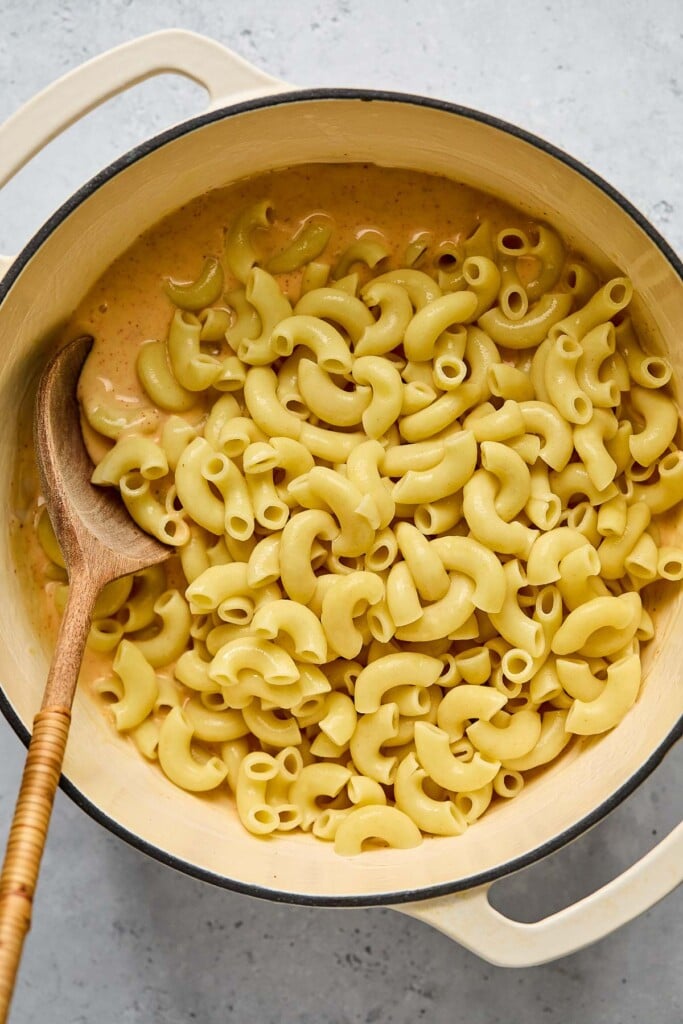 Large white dutch oven with cooked elbow pasta on top a cheese sauce before it is mixed together. There is a wooden spoon in the pot as well.