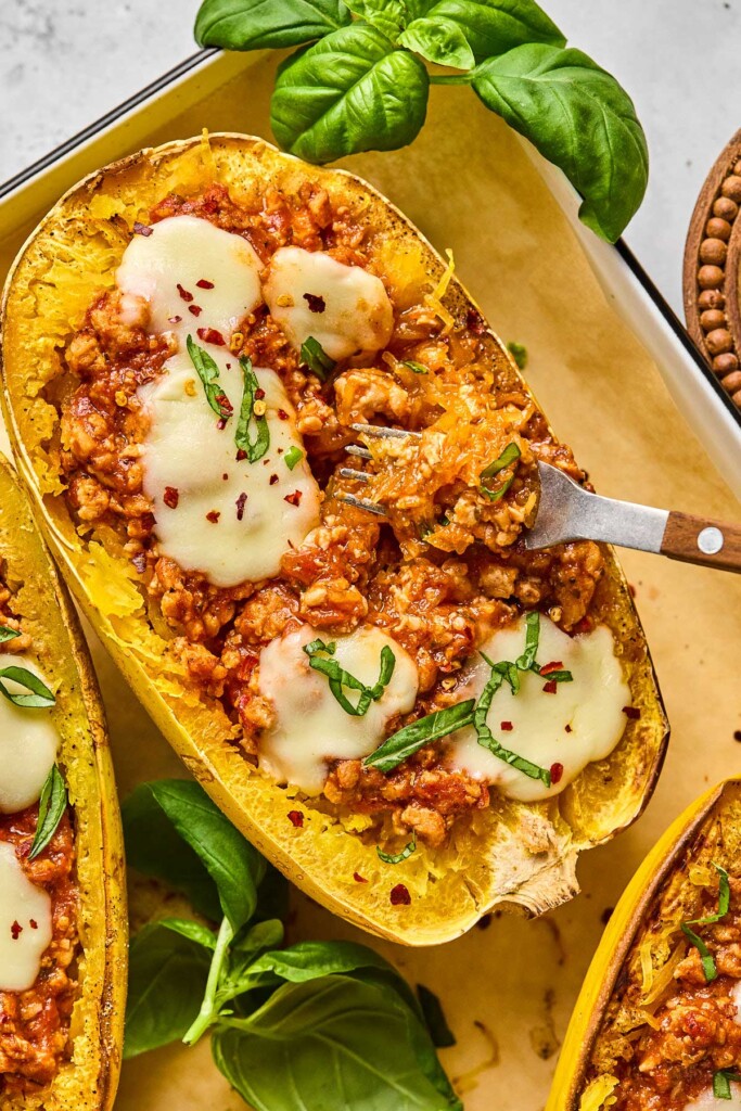 A close up of half a cooked spaghetti squash stuffed with meat sauce and topped with melted cheese, fresh basil, and red pepper flakes. A fork is in the spaghetti squash pulling out some of the noodles. Around it is more fresh basil.