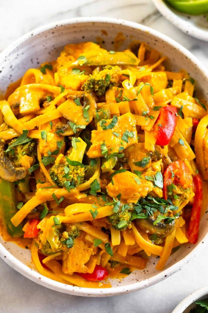 A close up of curry noodles with chicken and veggies in a white bowl topped with cilantro.