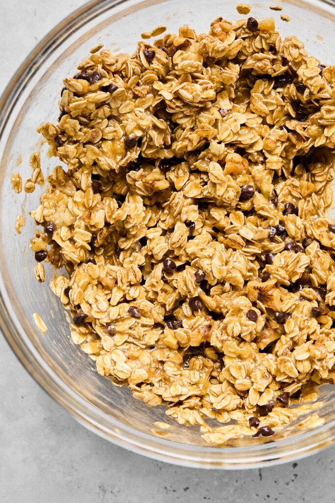Close up of a glass mixing bowl filled with a batter of mashed bananas, rolled oats, chocolate chips, and walnuts.