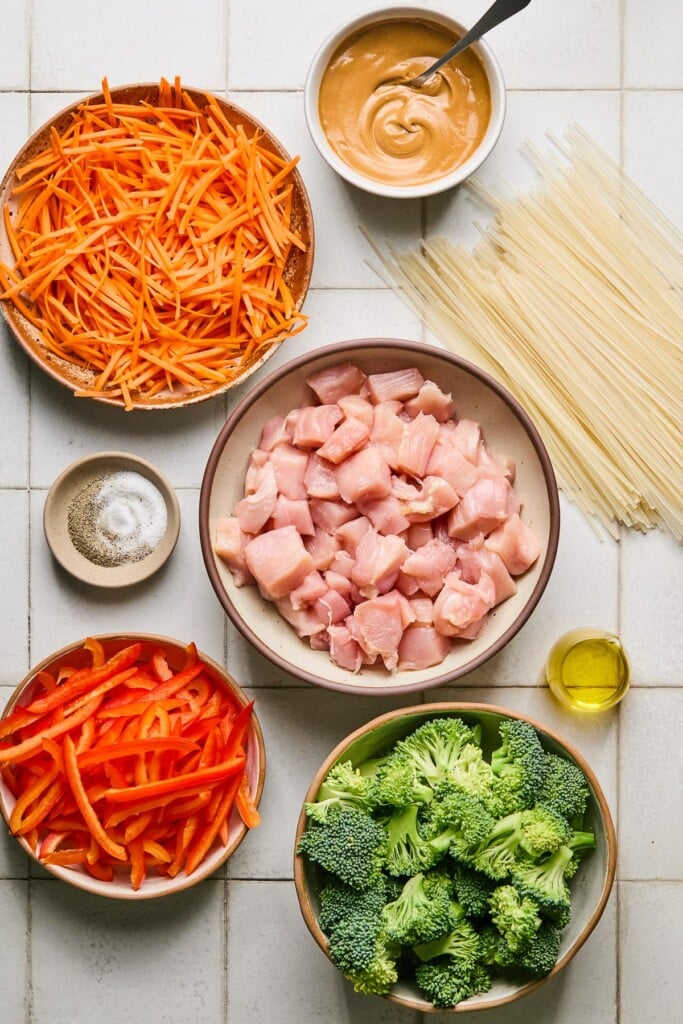 White tile counter with a dish of peanut butter sauce with a spoon in it, a pile of uncooked rice noodles, a bowl of cubed raw chicken, a small dish of olive oil, a bowl of broccoli florets, a bowl of julienned red bell pepper, a small dish of salt and pepper, and a bowl of julienned carrots.