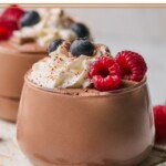 Two glass cups of protein pudding topped with whipped cream, fresh raspberries, fresh blueberries, and cinnamon. The cups are sitting on a white wooden board.