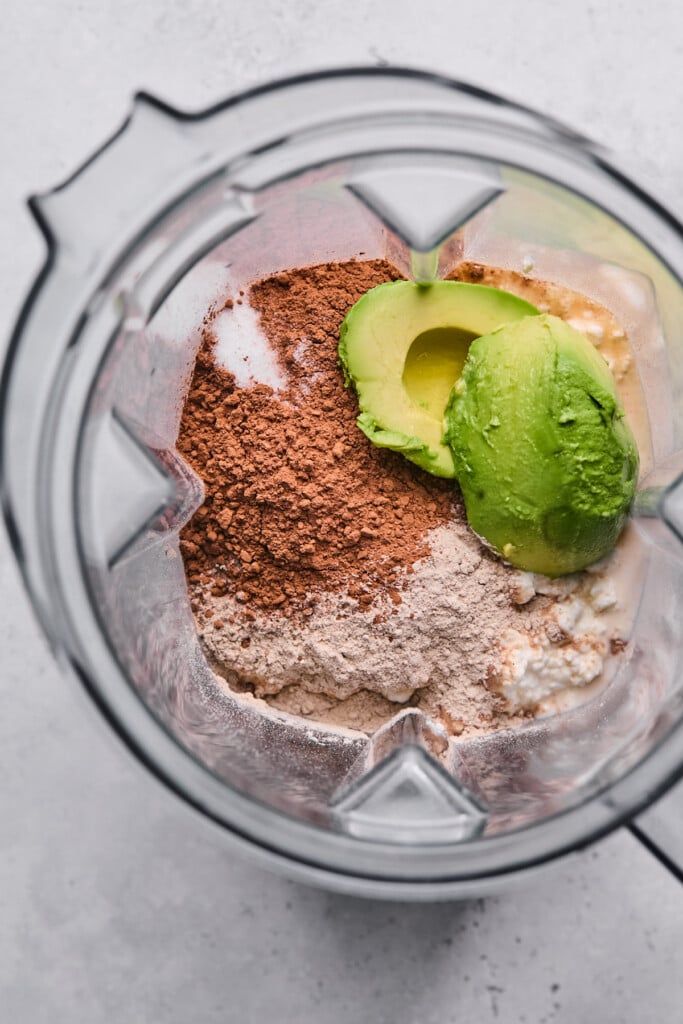 Blender filled with cottage cheese, cocoa powder, chocolate collagen, two halves of an avocado, and salt.