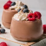 Two glass cups of protein pudding topped with whipped cream, fresh raspberries, fresh blueberries, and cinnamon. The cups are sitting on a white wooden board and there is more fresh fruit around them.