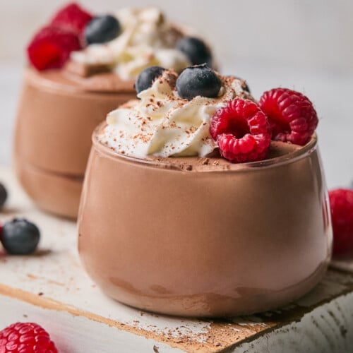 Two glass cups of protein pudding topped with whipped cream, fresh raspberries, fresh blueberries, and cinnamon. The cups are sitting on a white wooden board and there is more fresh fruit around them.