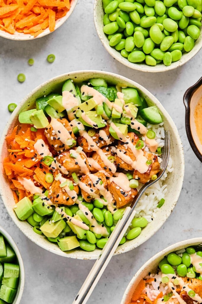 Salmon rice bowl with cubed salmon, white rice, edamame, diced avocado, shredded carrots, and diced cucumber with a fork in the bowl. Everything is drizzled with sriracha mayo and garnished with sesame seeds and green onions. Around the bowl is another rice bowl, a bowl of diced cucumber, a bowl of shredded carrots, a bowl of edamame, and a dish of sriracha mayo.