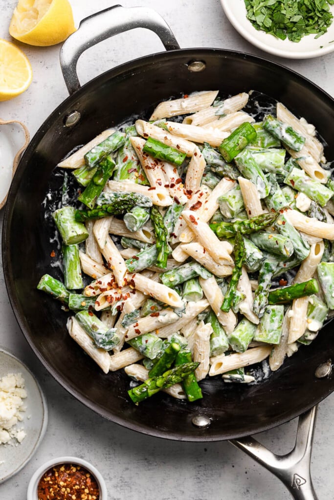 Creamy asparagus pasta tossed with a lemon feta sauce in a pan. it is topped with red pepper flakes. Around the pan is a bowl of herbs, a small dish of red pepper flakes, a dish of feta, and some lemons.