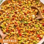 Buffalo chickpea salad with veggies, cheese, and herbs in a large bowl with two wooden spoons in it. Next to the bowl is fresh dill, half a lemon and juiced, and a jar of dressing.