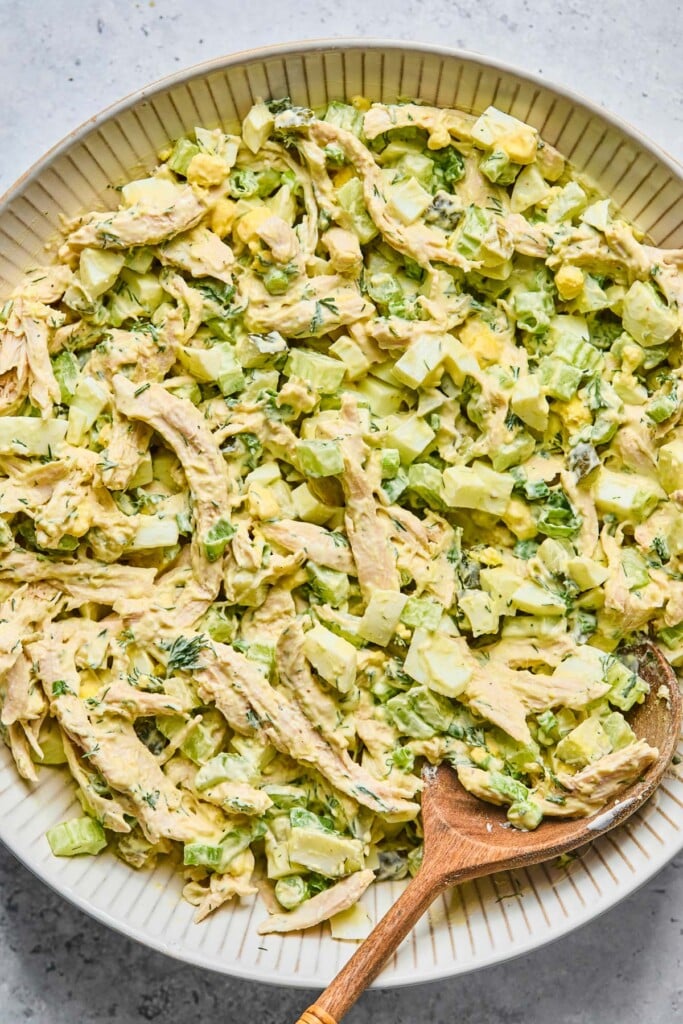 Chicken and egg salad with celery and dill in a large white bowl with a wooden spoon in it.