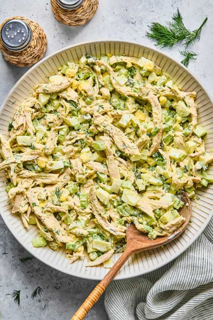 Overhead shot of chicken egg salad in a large white bowl with a wooden spoon in it. Around the bowl is a striped kitchen towel, a salt and pepper shaker, and some fresh dill.