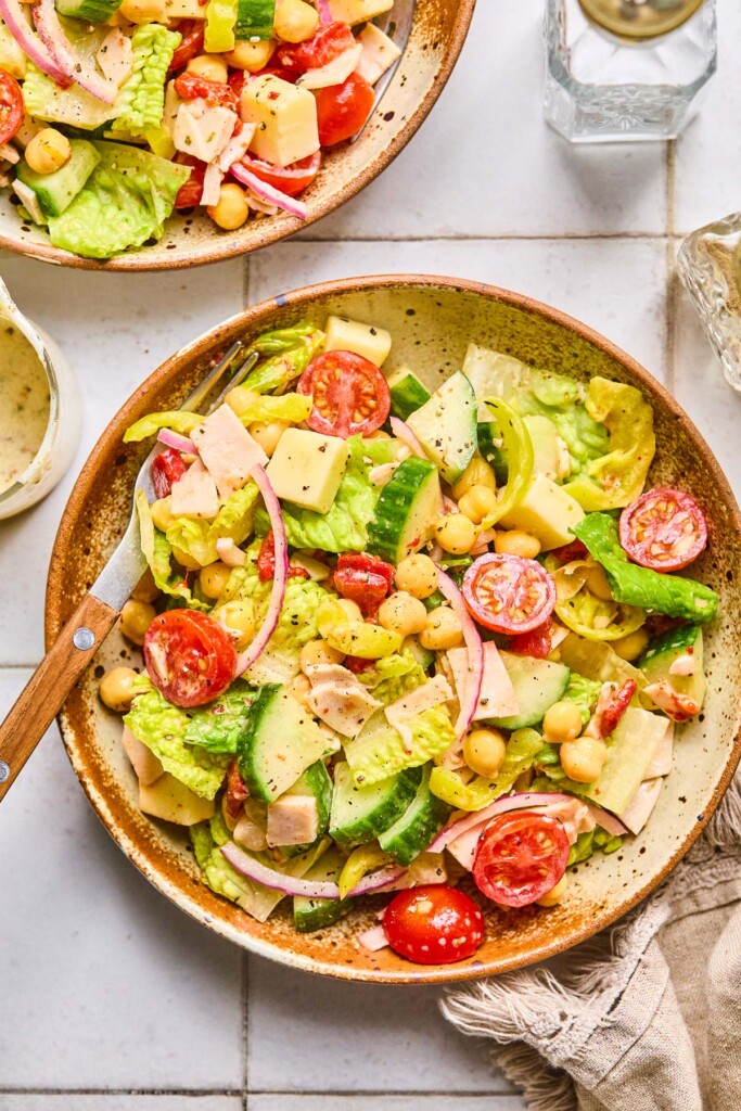 Overhead shot of a bowl of chopped salad with lettuce, tomato, cucumber, deli turkey, cheese, chickpeas, onions, and banana peppers with a fork in it. Next to the bowl is a jar of dressing, another bowl of salad, and a salt and pepper shaker.