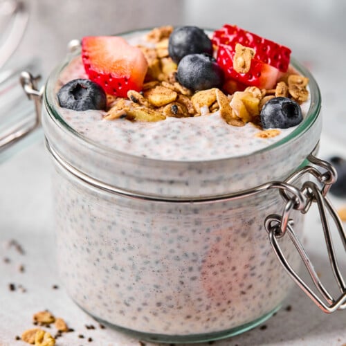 A jar of protein chia pudding topped with strawberries, blueberries, and granola. The jar is sitting on a plate and there is another jar filled with chia pudding behind the jar.