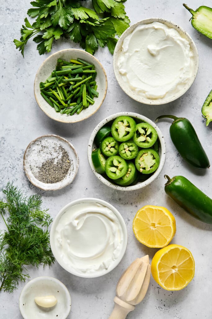 Grey concrete counter with a bowl of greek yogurt, a jalapeno cut in half, two whole jalapenos, a bowl of sliced jalapenos, a lemon that has been cut in half, a wooden citrus reamer, a bowl of sour cream, a small bowl with a clove of garlic, a bunch of fresh dill, a small bowl of salt and pepper, a bowl of chopped chives, and a bunch of parsley on it.