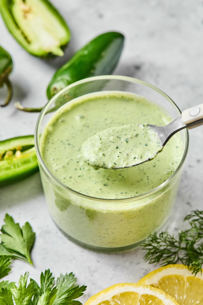 Jalapeno ranch dressing in a glass cup with a spoonful being held over it. Around the cup are fresh dill, some slices of lemon, fresh parsley, and some jalapenos.