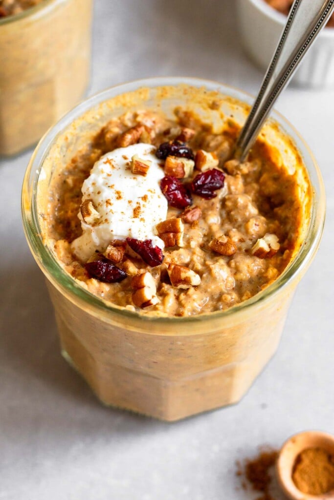 Two jars of pumpkin overnight oats, with one being behind the other. They are topped with greek yogurt, chopped pecans, dried cranberries, and cinnamon. A spoon is in the jar in the front. A ramekin of pecans is off to the side of the one in the back.