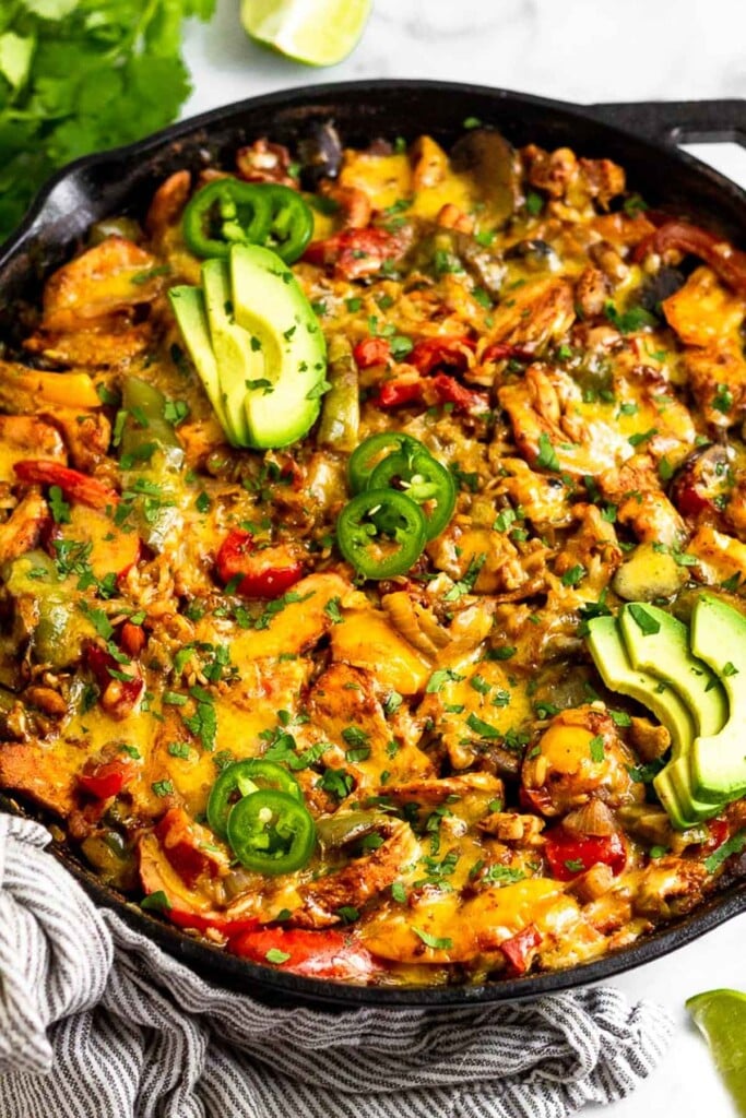 Large cast iron skillet with chicken fajita casserole with rice and topped with melted cheese. It is garnished with sliced avocado, jalapeños, and cilantro. Around the pan is more cilantro and lime wedges.