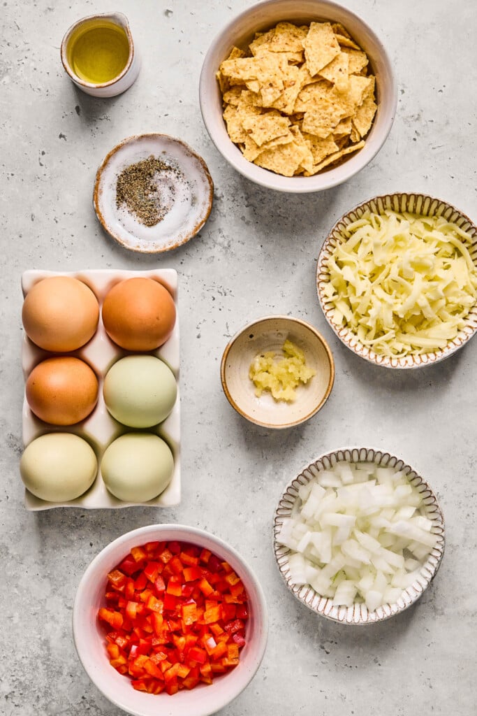 A bowl of broken tortilla chips, a bowl of shredded cheese, a bowl of minced garlic, a bowl of diced yellow onion, a bowl of diced red bell pepper, a half carton of eggs, a bowl of salt and pepper, and a small cup of olive oil on a grey concrete countertop.