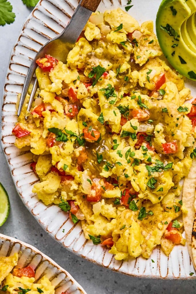 Mexican migas garnished with cilantro and salsa on a plate. Also on the plate is a fork and some sliced avocado. Next to the plate is another plate of migas.