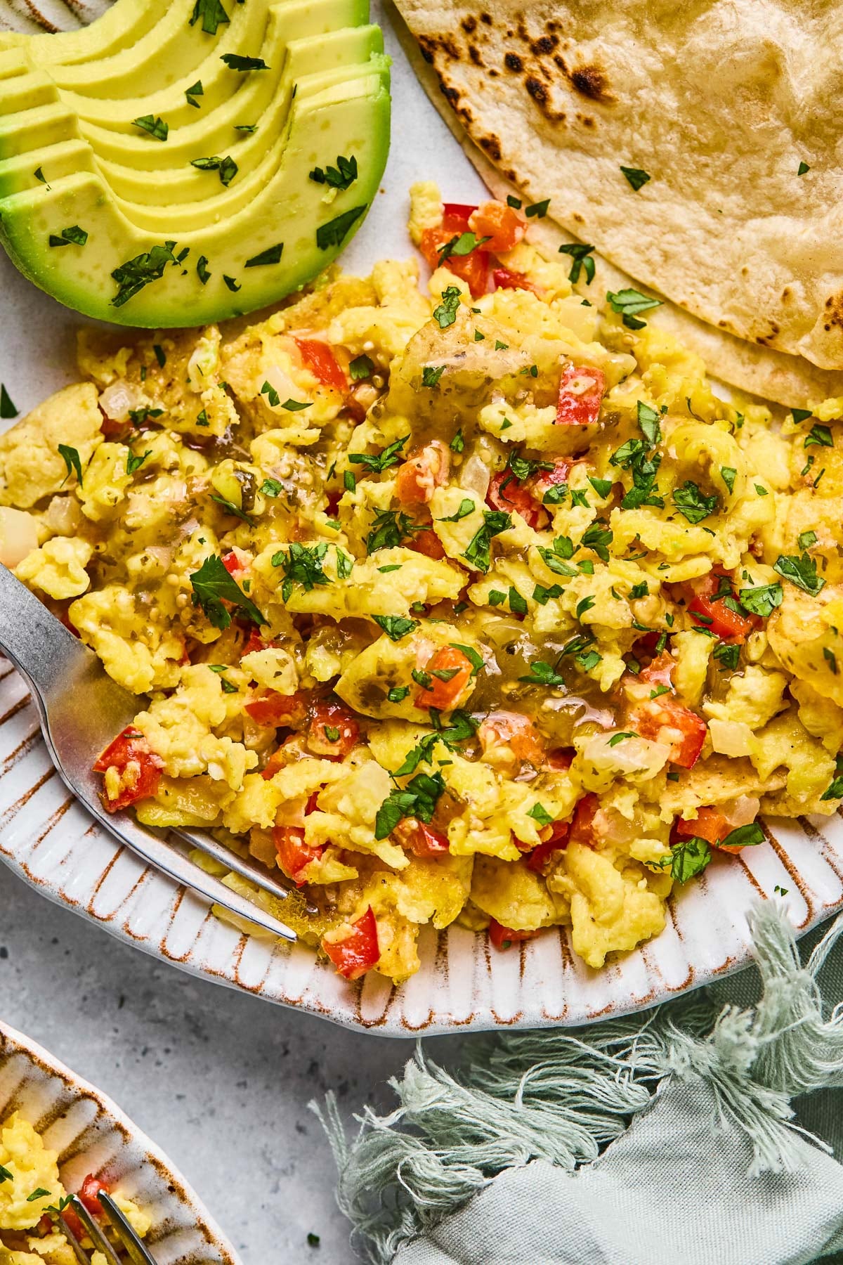A plate of migas, sliced avocado, and tortillas. The migas are garnished with salsa and cilantro. Next to the plate is a green kitchen towel and another plate of migas.