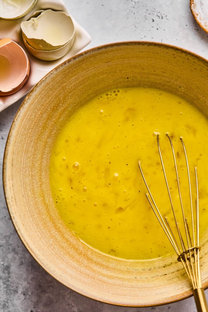 Whisked eggs in a bowl with a whisk in it. Next to the bowl is a carton with egg shells in it.