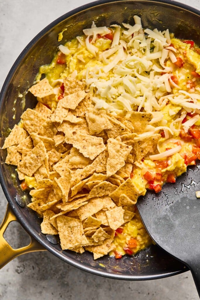 Large skillet with eggs that are being scrambled with onions and peppers. Overtop the partially scrambled eggs is some shredded cheese and broken tortilla chips before they are mixed in. A spatula is off to the side in the skillet.