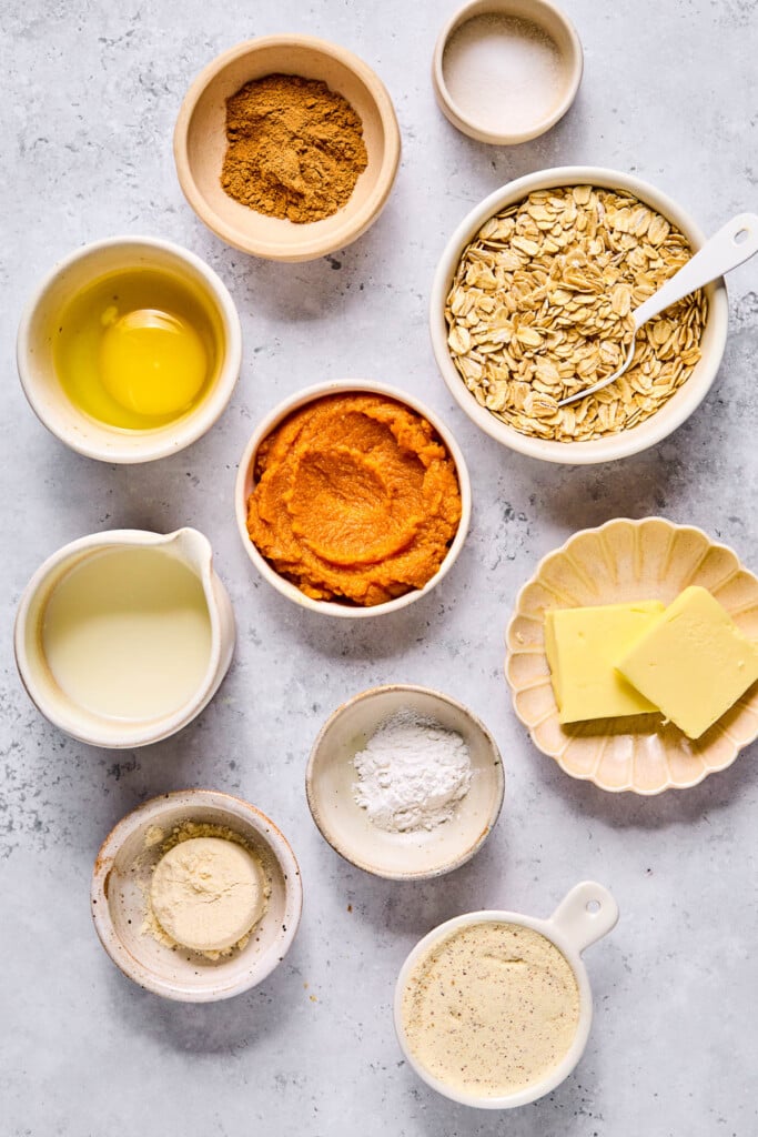 A small dish of salt, a bowl of rolled oats with a spoon in it, a plate with two slices of butter, a bowl of baking powder, a bowl of vanilla protein powder, a small bowl of coconut flour, a small pitcher of milk, a bowl of pumpkin puree, a bowl with a cracked egg in it, and a small bowl of pumpkin spice on a grey concrete counter.
