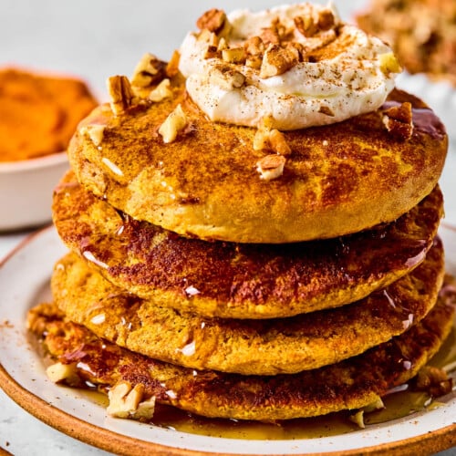 A stack of pumpkin protein pancakes topped with greek yogurt, pecan pieces, and maple syrup on a plate. Around the plate are some pecan pieces, cinnamon sticks, a bowl of pumpkin puree, and bowl of pecans.