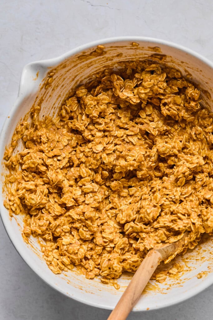 White mixing bowl with a wet batter with rolled oats in it. A wooden spoon is also in the bowl.