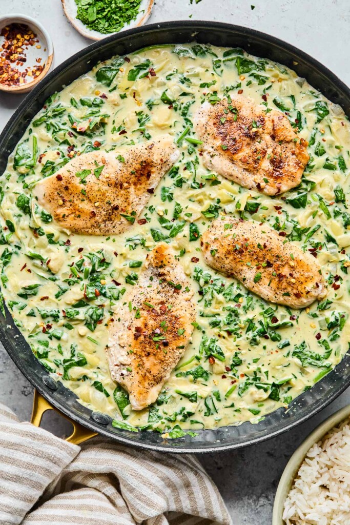 Spinach artichoke chicken skillet garnished with red pepper flakes. Next to the pan is a bowl of rice, a striped kitchen towel, a small bowl of red pepper flakes, and a small bowl of parsley.