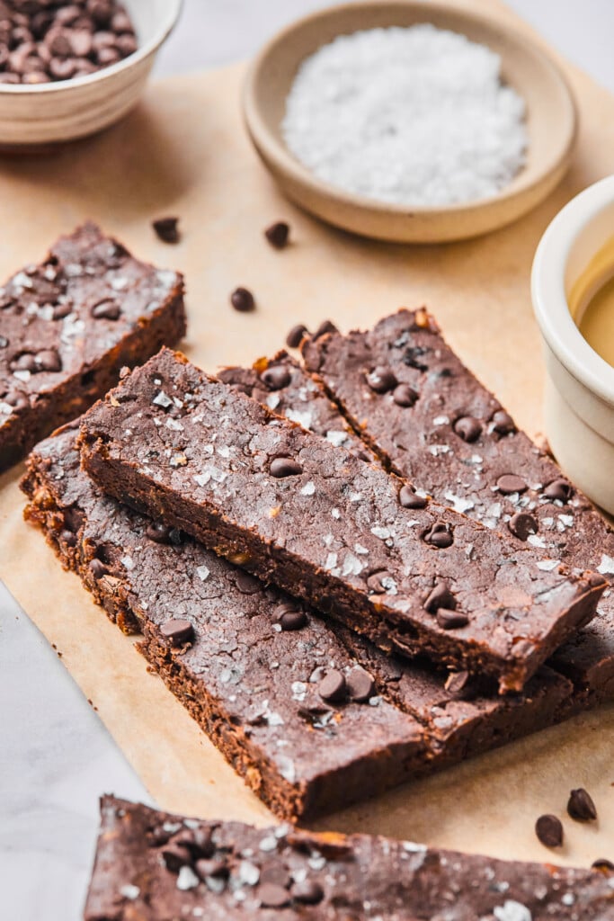 Three dark chocolate protein bars lined up next to each other with one on top of them all. The bars are on top of a piece of brown parchment paper and around them are some more protein bars, some mini chocolate chips, a bowl of flaky sea salt, and a bowl of mini chocolate chips.