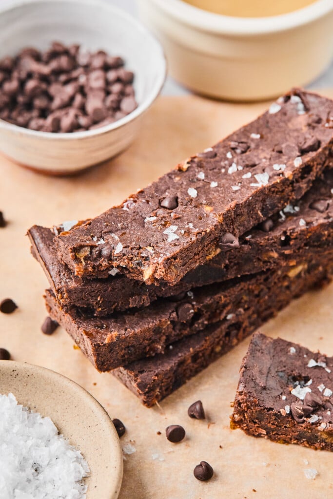 Four chocolate protein bars stacked on top of each other. The one on the top has a bite taken out of it. Around the stack are some mini chocolate chips, another protein bar, a bowl of flaky sea salt, a bowl of mini chocolate chips, and a bowl of tahini.