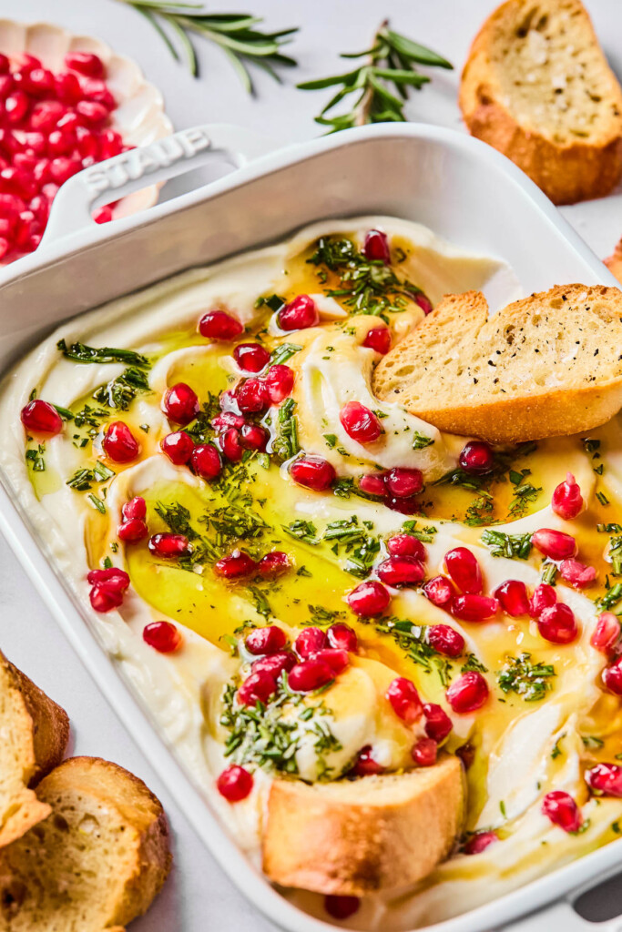 A dish of whipped ricotta with honey, rosemary, olive oil, and pomegranates on top of it. Two pieces of crostini are dipped into it. Around the dish are more crostini, a bowl of pomegranate seeds, and fresh rosemary sprigs.