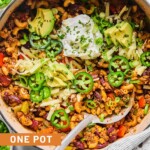 Overhead shot of chili pasta in a large Dutch oven. It is topped with sour cream, shredded cheese, sliced avocado, sliced jalapeños, sliced green onions, and cilantro. A metal spoon is also in the pot. Around the pot is a striped kitchen towel, a bowl of sliced green onions, a bowl of sliced jalapeños, a bowl of shredded cheese, and a bowl of chopped cilantro.