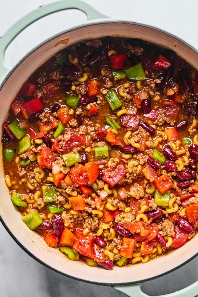 Large Dutch oven filled with a mixture of cooked ground beef, diced peppers, uncooked pasta, tomato sauce, and broth before it is cooked.