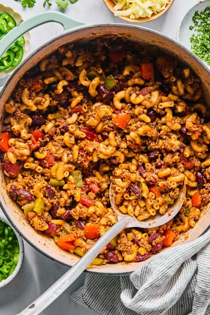 Overhead shot of chili pasta in a large Dutch oven with a metal spoon in it. Around the pot is a striped kitchen towel, a bowl of sliced green onions, a bowl of sliced jalapeños, a bowl of shredded cheese, and a bowl of chopped cilantro.