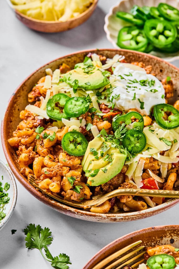 Chili with pasta topped with sour cream, shredded cheese, sliced avocado, sliced jalapeños, and cilantro in a bowl. A gold fork is also in the bowl. Around the bowl is another bowl of chili, some cilantro leaves, a bowl of shredded cheese, and a bowl of sliced jalapeños.