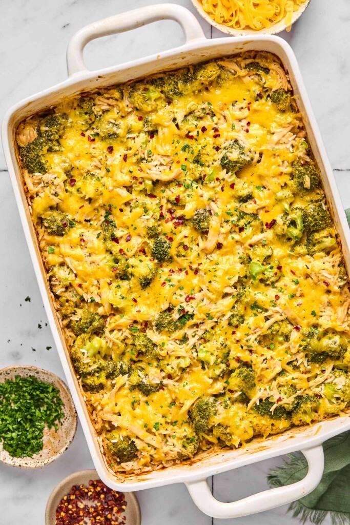 Healthy chicken broccoli rice casserole covered with melted cheese and topped with red pepper flakes and fresh parsley in a large baking dish. Next to the dish is a small bowl of red pepper flakes, a small bowl of parsley, and a bowl of shredded cheese.
