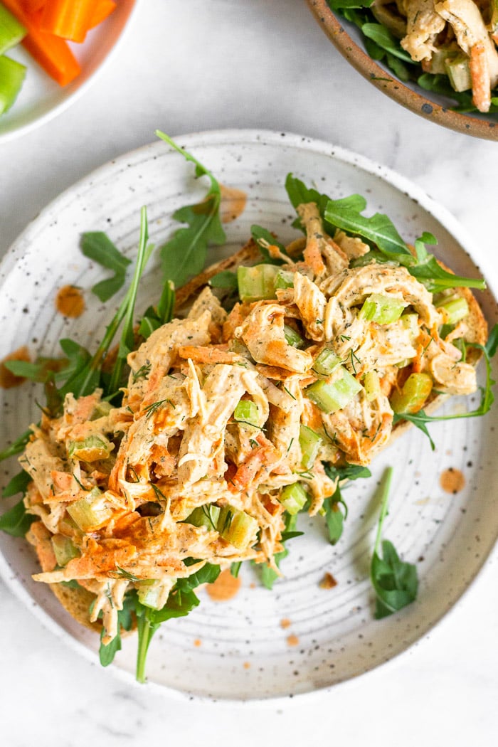 Healthy chicken salad with celery, carrots, and buffalo sauce on top of some arugula on a piece of bread on a plate. It is drizzled with hot sauce. Around the plate is a plate of carrots and celery and a bowl of more chicken salad.