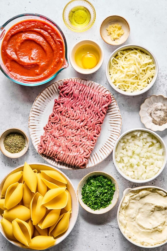 A small bowl of olive oil, a small bowl of minced garlic, a small dish with a cracked raw egg in it, a bowl of shredded mozzarella cheese, a dish with salt and pepper in it, a bowl of diced onions, a bowl of ricotta cheese, a bowl of chopped parsley, a bowl of uncooked jumbo pasta shells, a small bowl with Italian seasoning, a plate with a pound of raw ground beef, and a large measuring cup of pasta sauce on a grey counter.