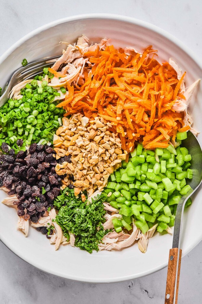 A large white bowl with shredded chicken topped with a pile of shredded carrots, a pile of diced celery, a pile of chopped cilantro, a pile of raisins, a pile of chopped green onions, and a pile of chopped roasted cashews before it is mixed together. A spoon is in the bowl as well.