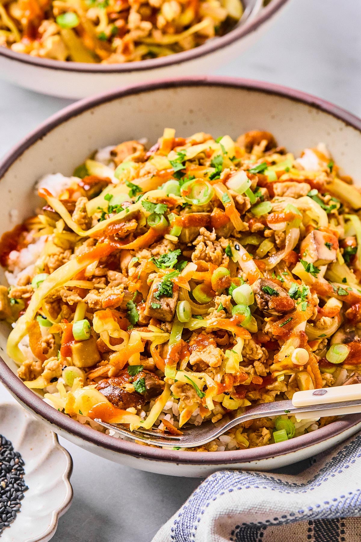 Healthy egg roll in a bowl with ground chicken, sliced cabbage, shredded carrots, and mushrooms over white rice. It is in a bowl and garnished with sriracha, green onions, cilantro, and sesame seeds. A fork is also in the bowl. Around the bowl is a kitchen towel, a bowl of black sesame seeds, and another egg roll bowl.