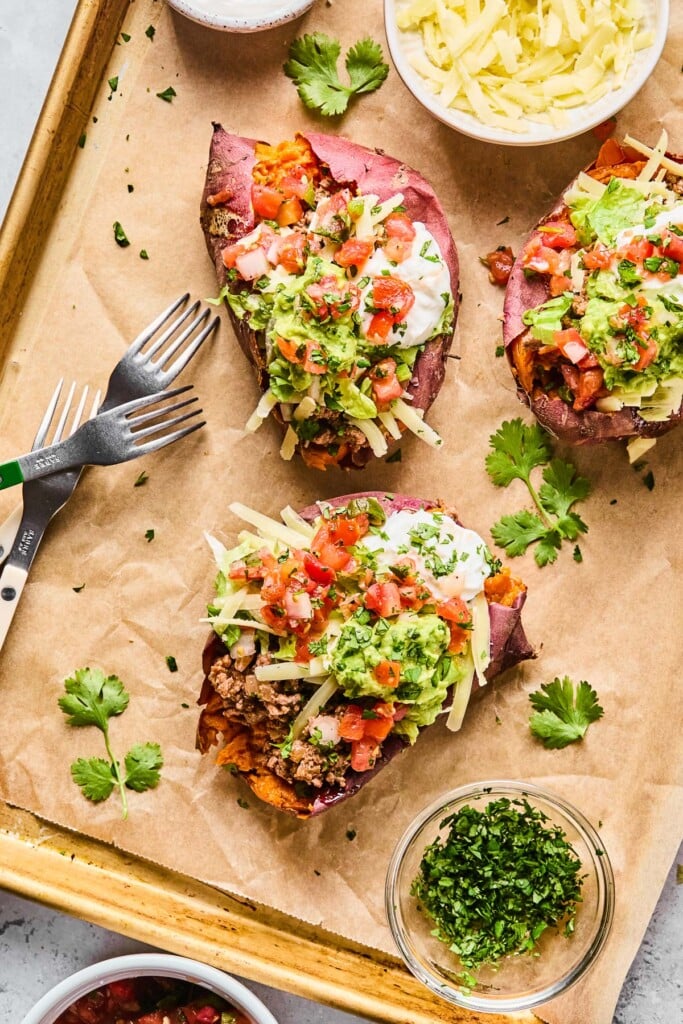 Overhead shot of three taco stuffed sweet potatoes on a baking sheet lined with parchment paper. They are topped with shredded cheese, guacamole, pico de gallo, greek yogurt, and cilantro. Also on the baking sheet are two forks, a bowl of shredded cheese, cilantro leaves, and a bowl of chopped cilantro.