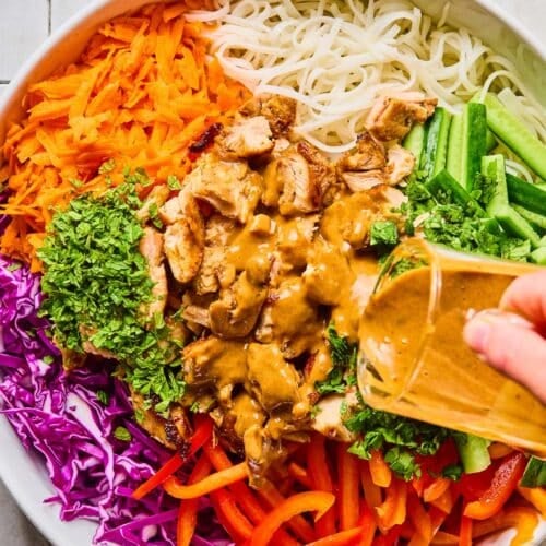 A large bowl of cold noodle salad with piles of rice noodles, cucumber, red bell pepper, cabbage, carrots, seasoned chicken, and fresh herbs with a creamy almond butter dressing being poured over top of it before it is all mixed.