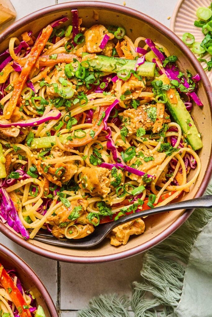 Spring roll in a bowl with chicken, veggies, rice noodles, and fresh herbs in a creamy brown sauce. It is topped with sliced scallions, fresh herbs, and sesame seeds. There is also a fork in the bowl. Around the bowl is a green kitchen towel, another bowl of noodle salad, and a plate of chopped scallions.