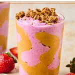 Peanut butter and jelly smoothie in a glass topped with granola. The glass has some peanut butter swirled inside it. Around the glass are some fresh strawberries, a bowl of peanut butter, and another glass with a smoothie in it.