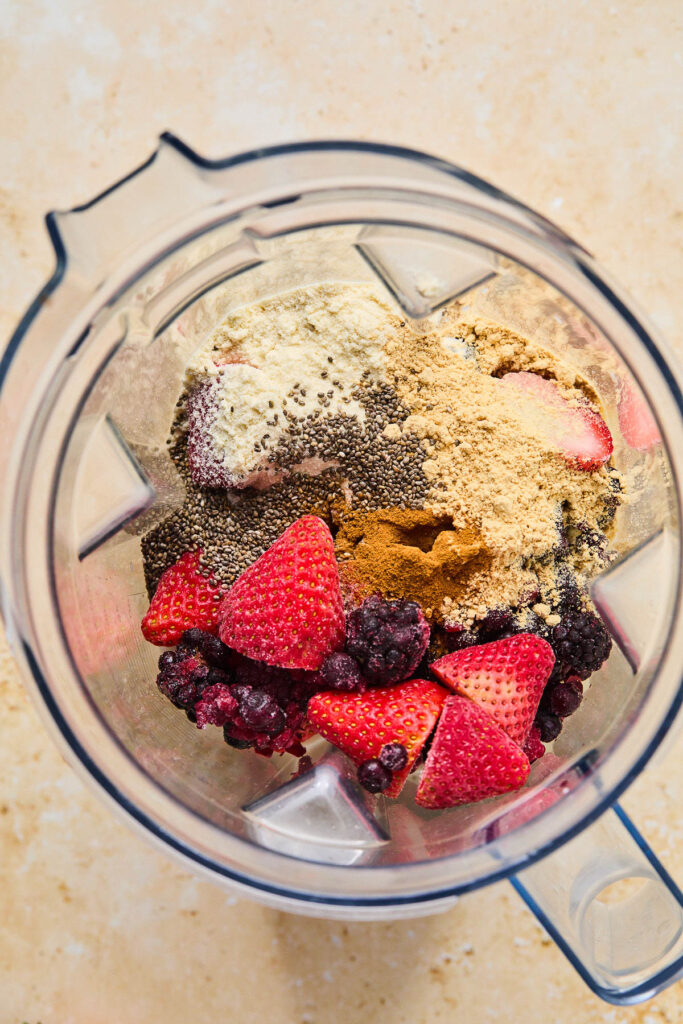 Overhead shot of a blender filled with frozen berries, chia seeds, protein powder, peanut butter powder, and cinnamon before it is blended.